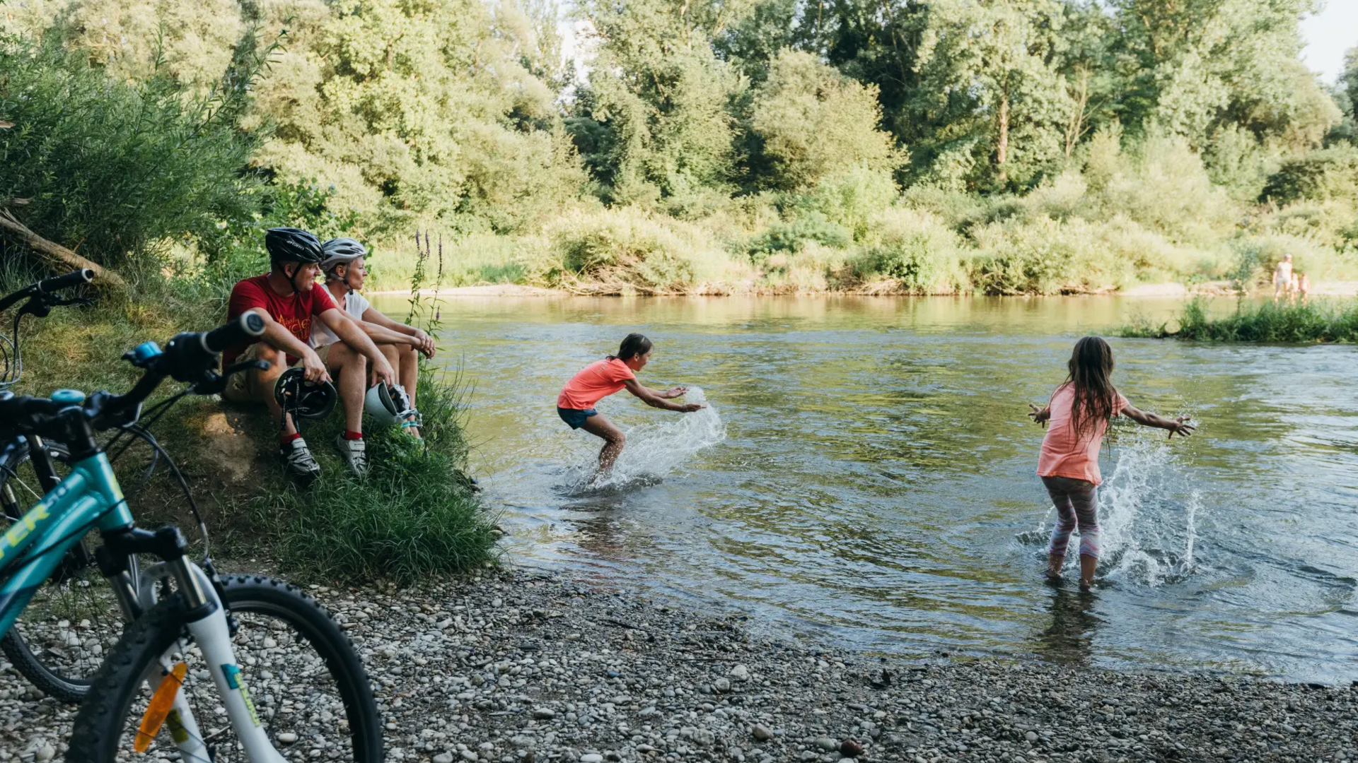 Children splashing in a river as two men sit on riverbank with bikes nearby