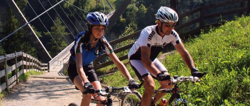 Two cyclists on forest path near suspension bridge and green hills