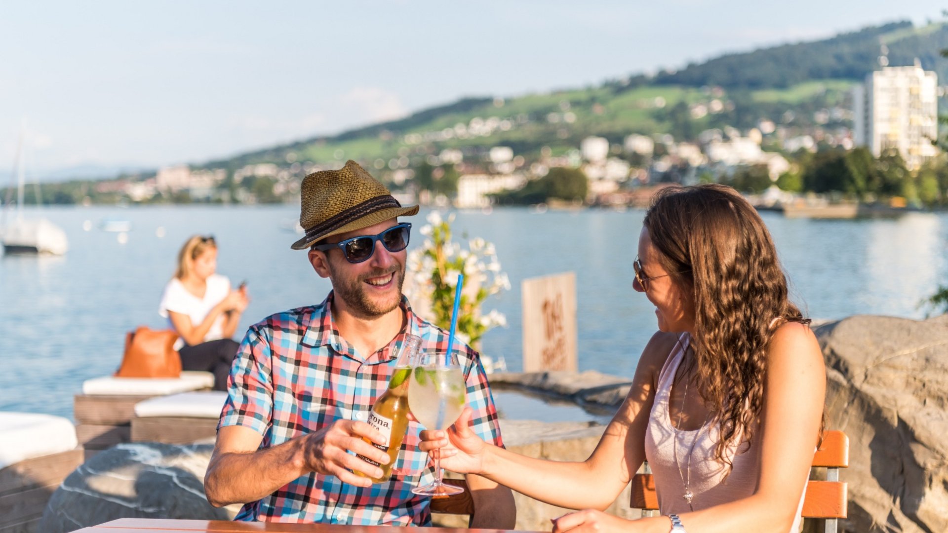 Couple clinking drinks at lakeside outdoor seating on sunny day