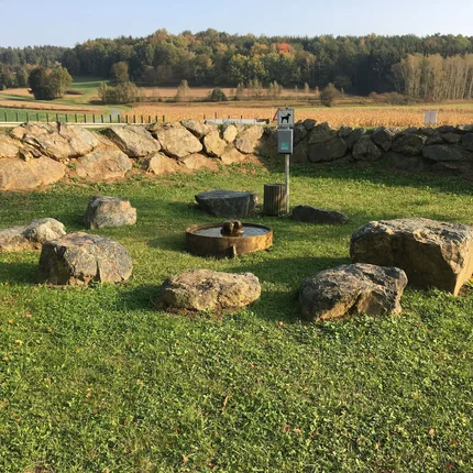 Runder Steinkreis mit Wasserbrunnen auf Gras vor einer Steinmauer und Wald