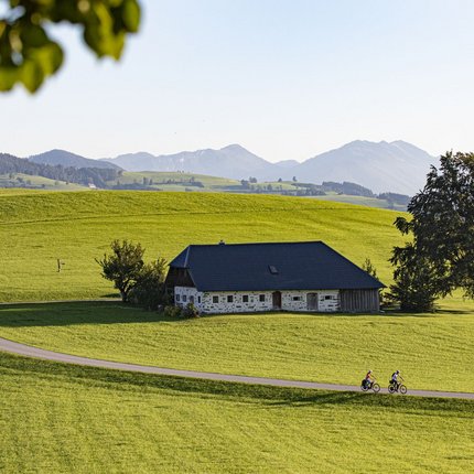 Cyclists on country road by house on green hill with mountains in background