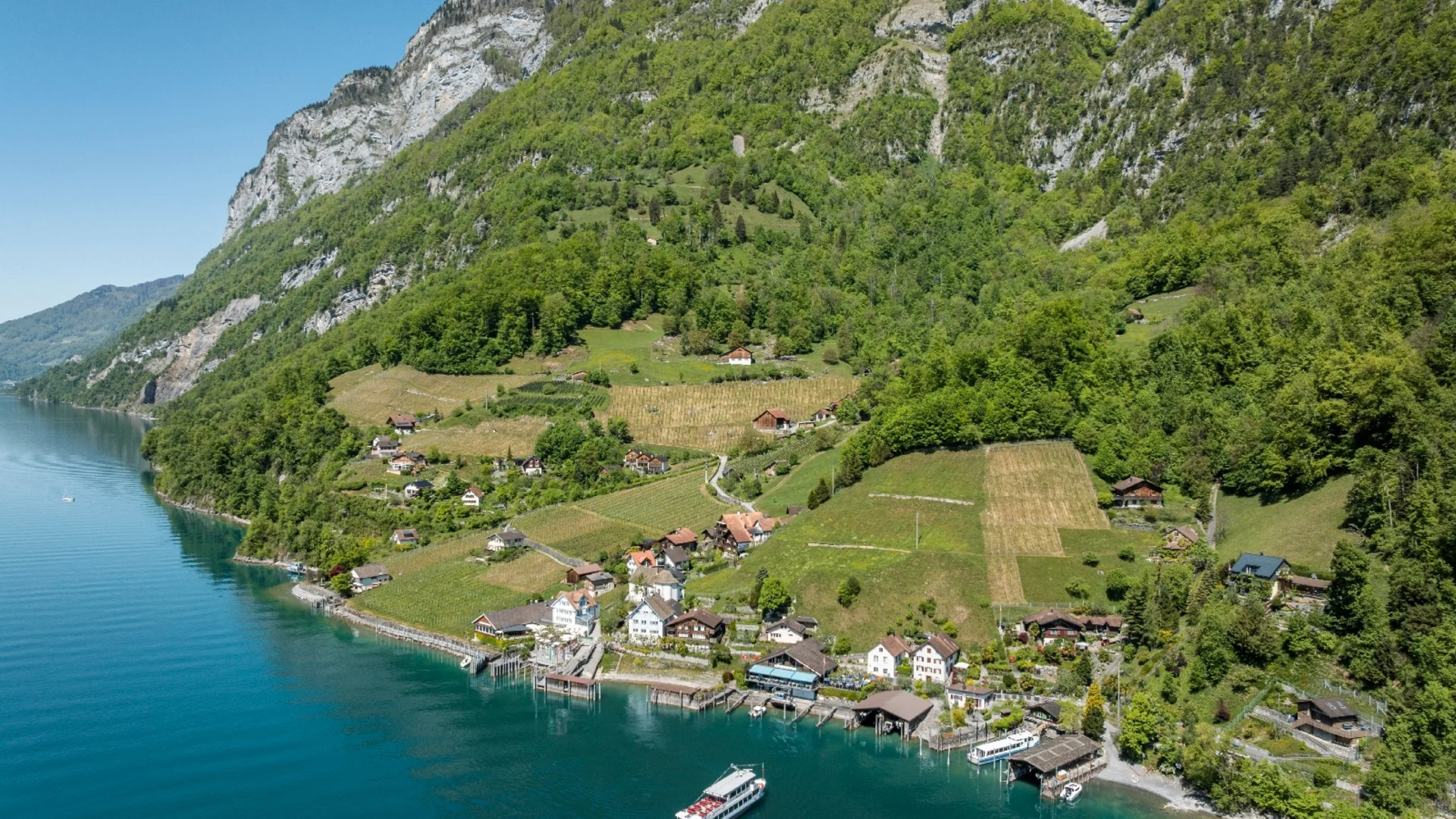 Dorf am Seeufer vor bewaldetem Berg unter klarem Himmel
