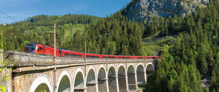 Roter Zug fährt über Bogenbrücke im grünen Bergwald bei Sonnenschein