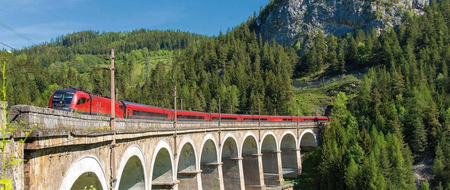 Red train crossing arched bridge in green forested mountains under clear sky