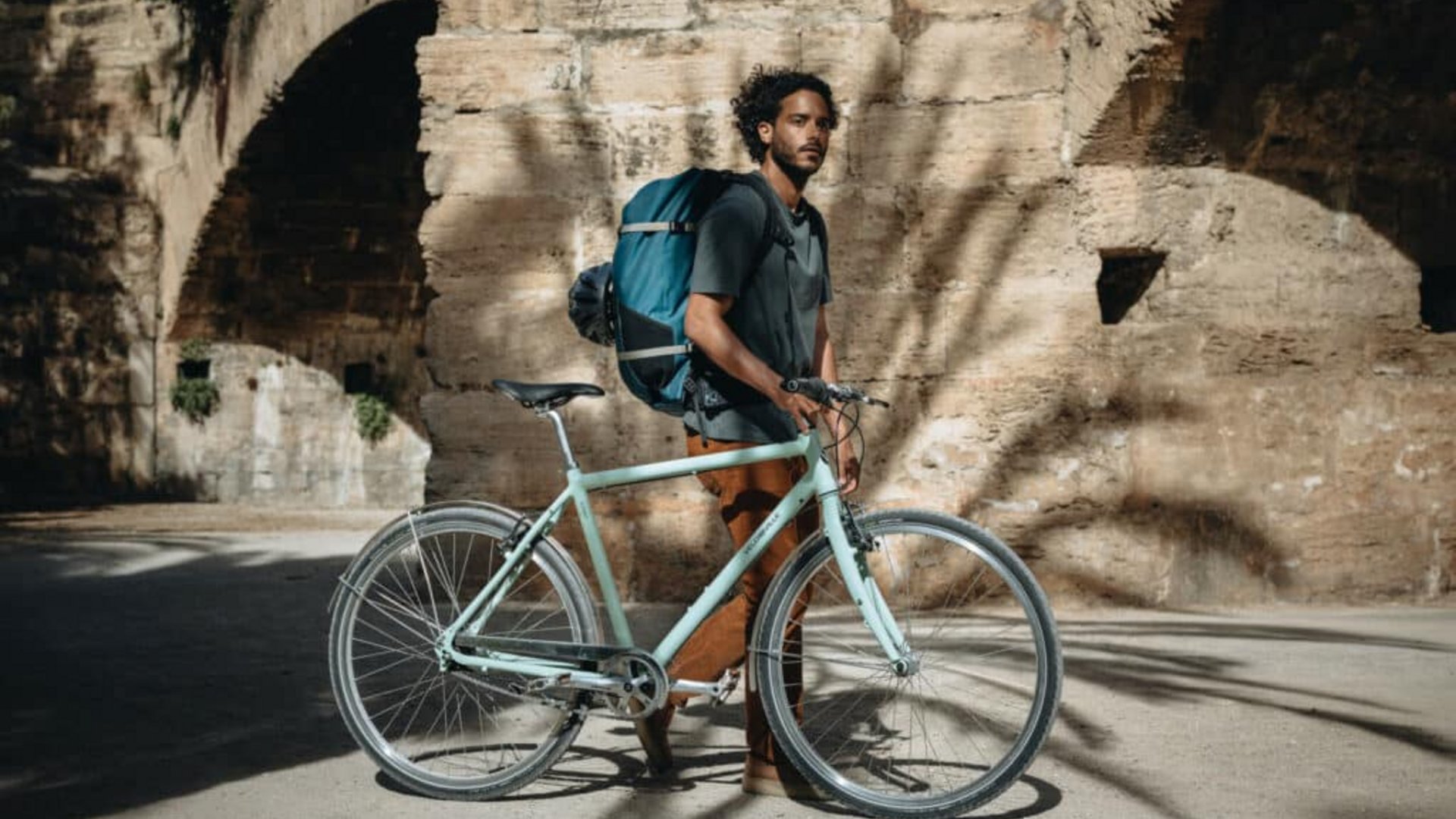 Man with backpack standing next to bike by stone wall