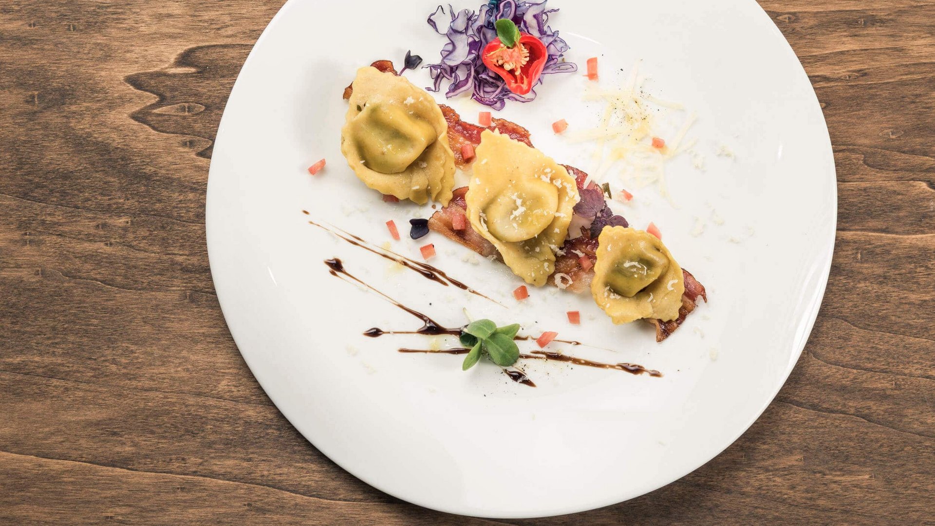 Three ravioli on a white plate with vegetables and balsamic decoration