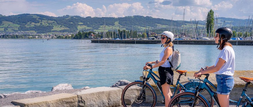 Two women with helmets and bikes by the lake with mountains in the background