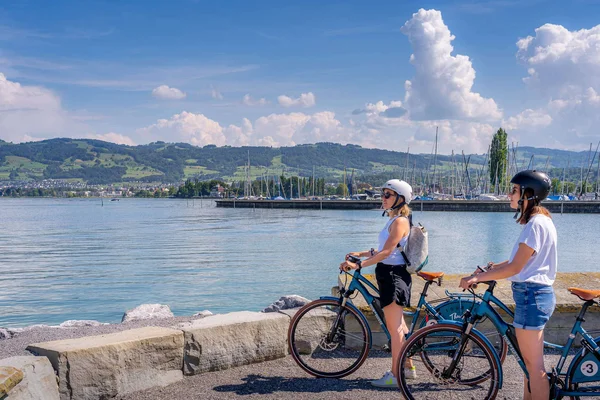 RheinWelten – Eine Reise entlang des Wassers © Thomas Staub Zwei Frauen mit Helmen und Fahrrädern am See mit Bergen im Hintergrund