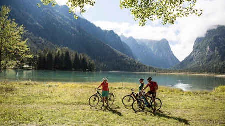 Radreisen in der Gruppe © Velontour Drei Radfahrer an einem Bergsee mit bewaldeten Bergen im Hintergrund
