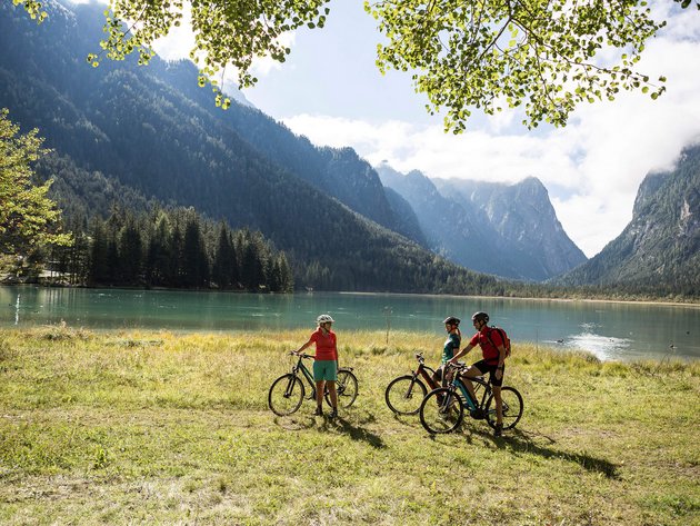 Drei Radfahrer an einem Bergsee mit bewaldeten Bergen im Hintergrund
