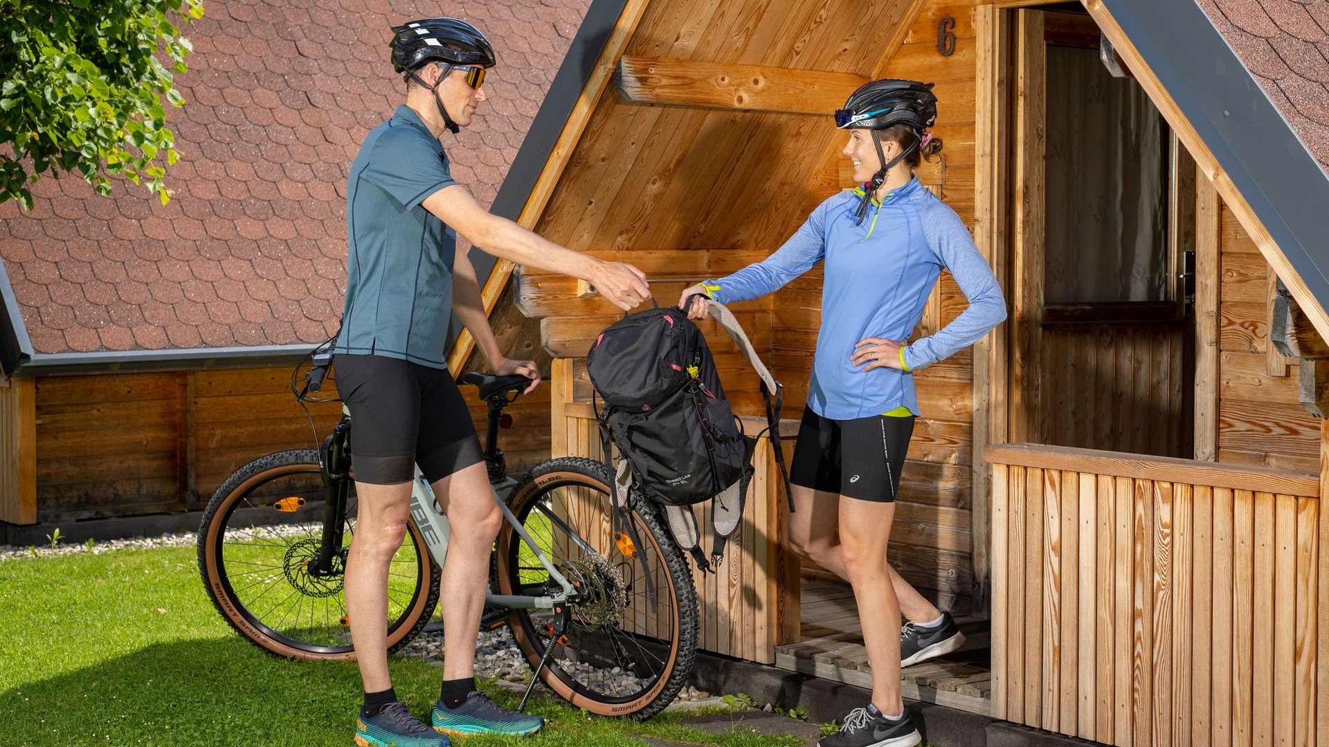 Two cyclists exchanging backpack by wooden cabin