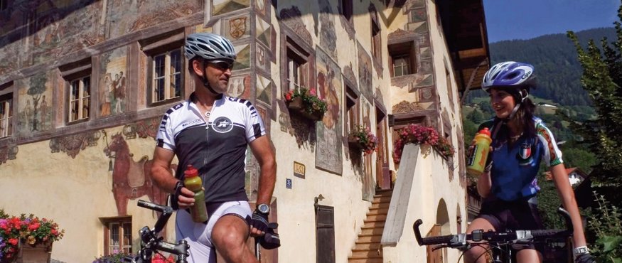 Two cyclists with helmets drinking water in front of a painted house