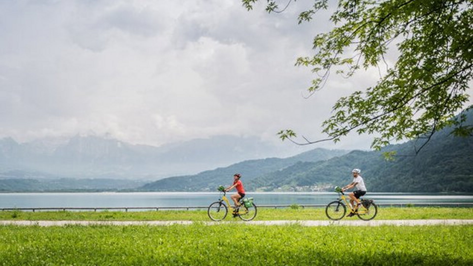 Two people cycling along lakeside with mountains and cloudy sky behind