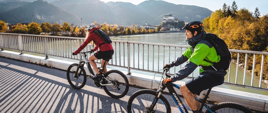 Two cyclists riding on a bridge with mountains and river in the background