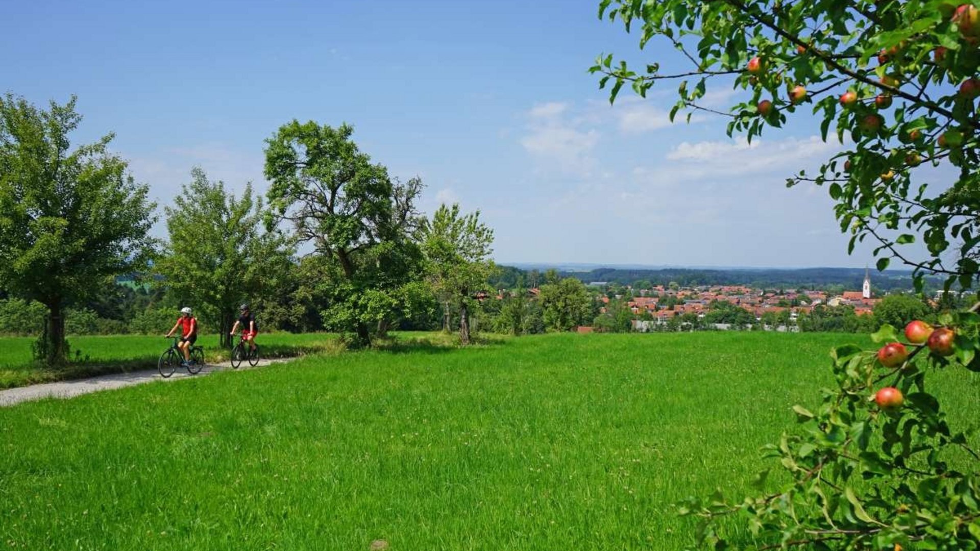 Two cyclists on a path in green fields with a village in the distance