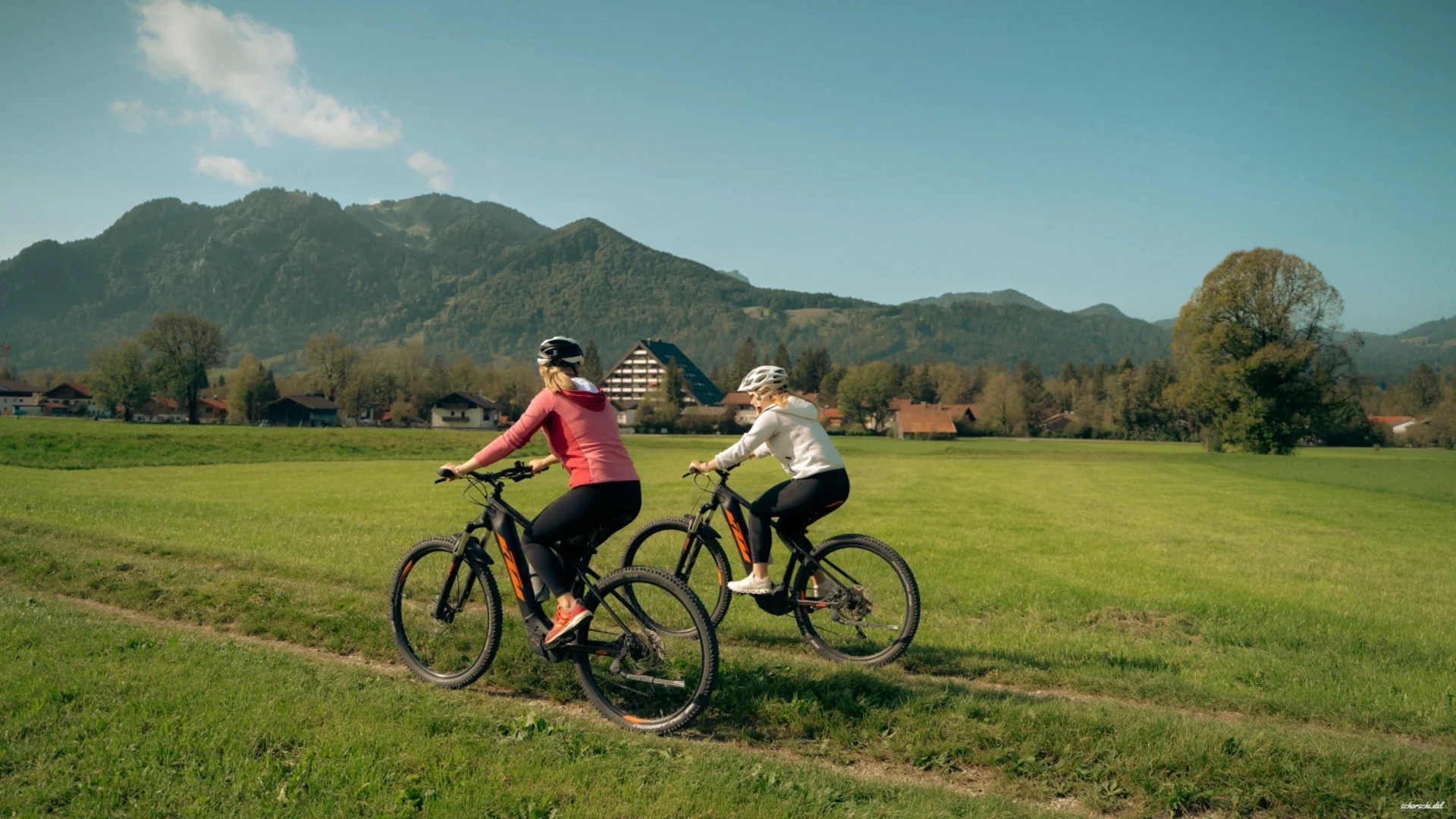 Zwei Radfahrerinnen fahren auf einem Feldweg mit Bergen im Hintergrund
