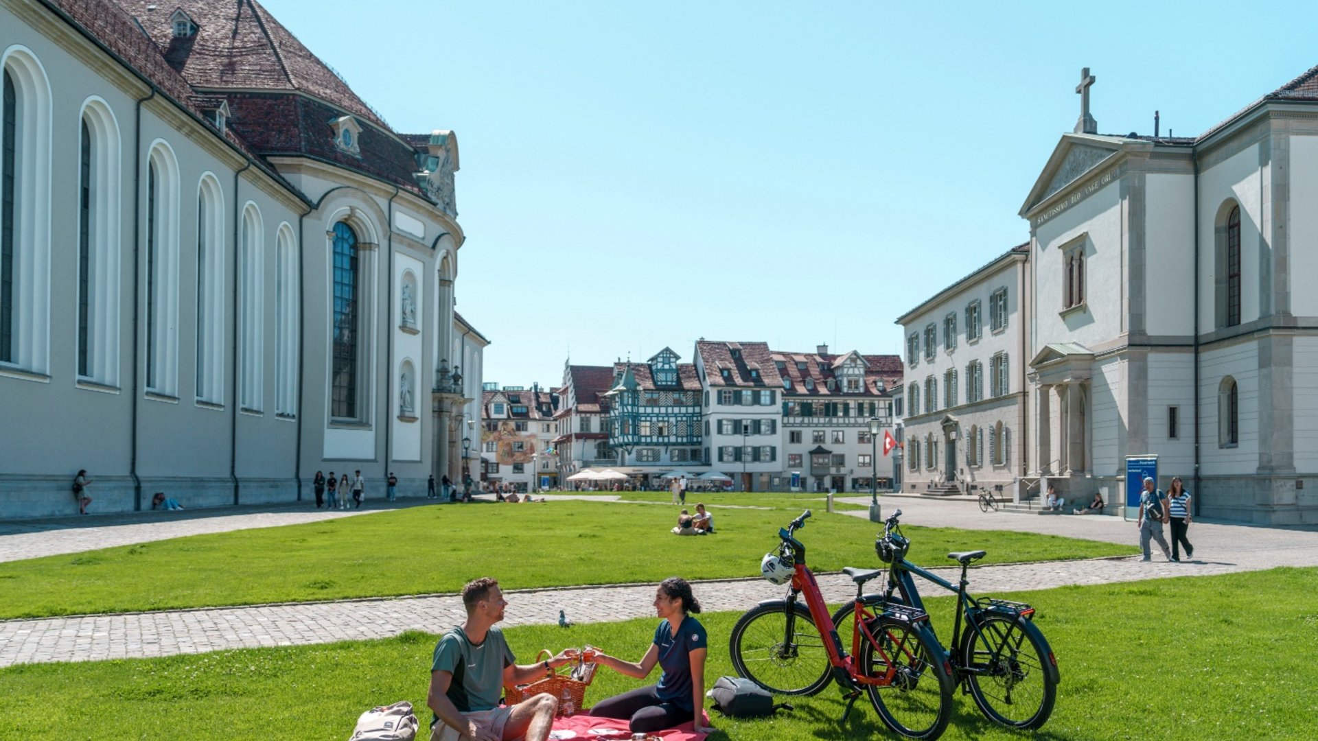 People picnic on green lawn in front of historic buildings on a sunny day