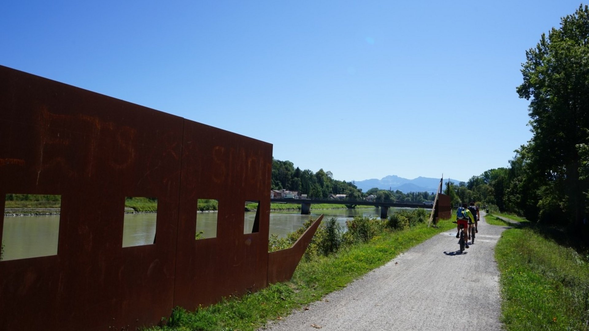 Cyclists on a path by a river with metal art and a bridge in the distance
