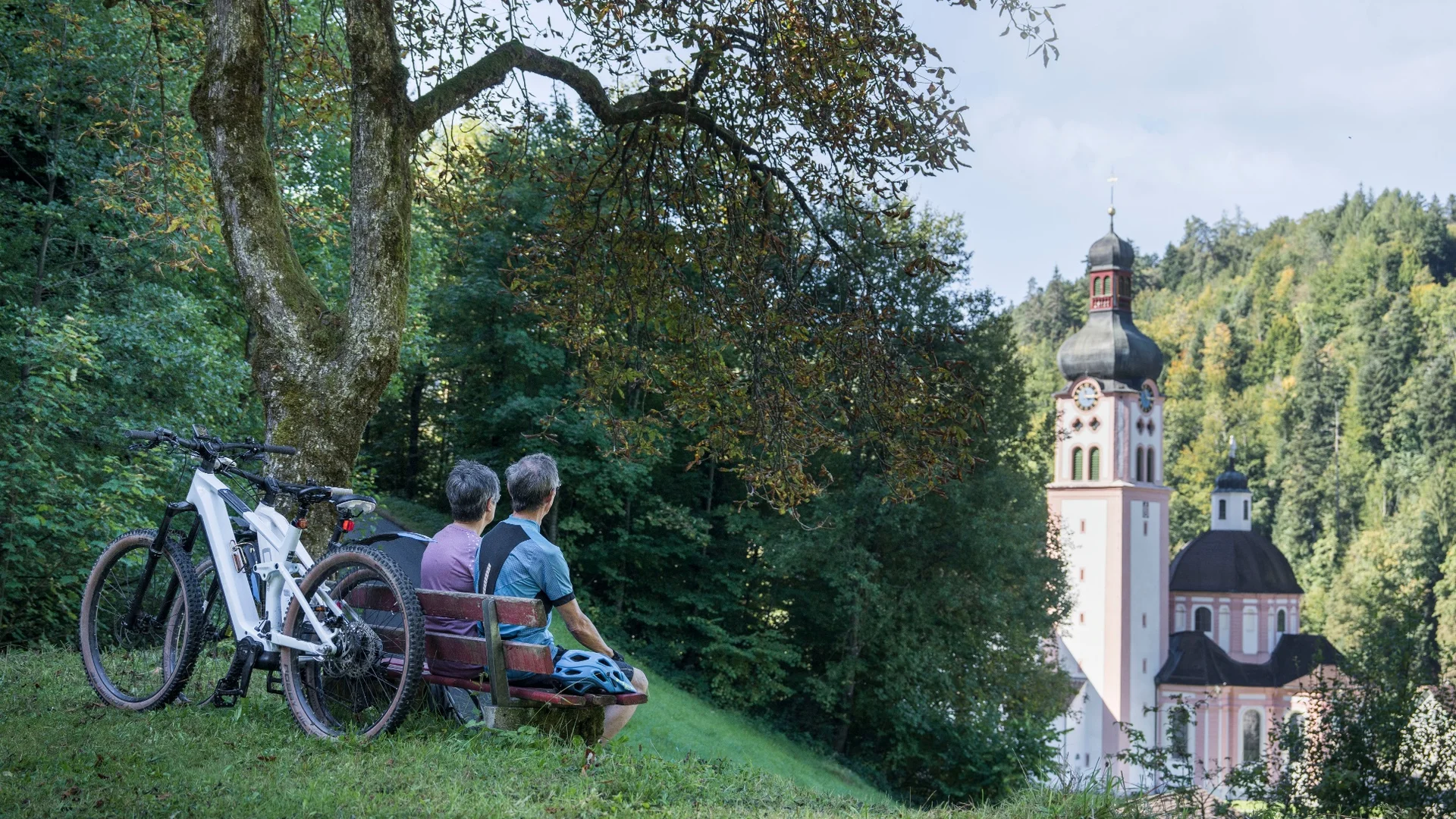 Zwei Radfahrer sitzen auf einer Bank und blicken auf eine Kirche im Wald.