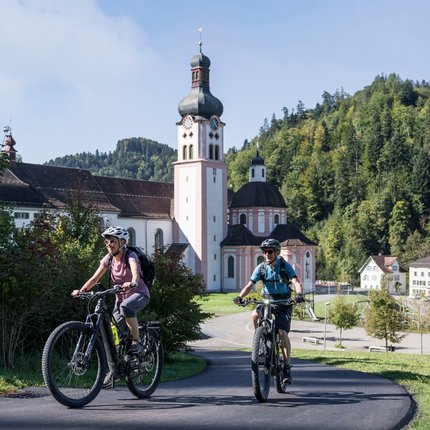 Two cyclists riding near a church and forest in a rural area
