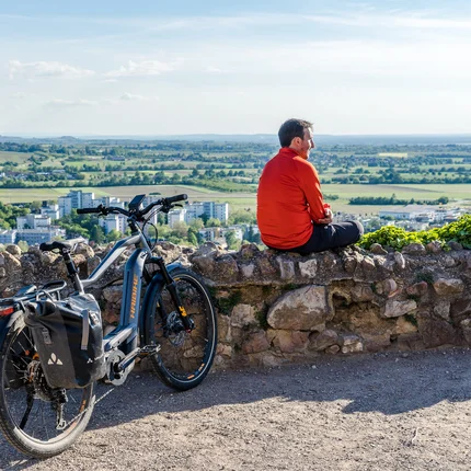 Mann in rotem Jacke sitzt auf Steinmauer mit E-Bike und Aussicht auf Stadt und Landschaft