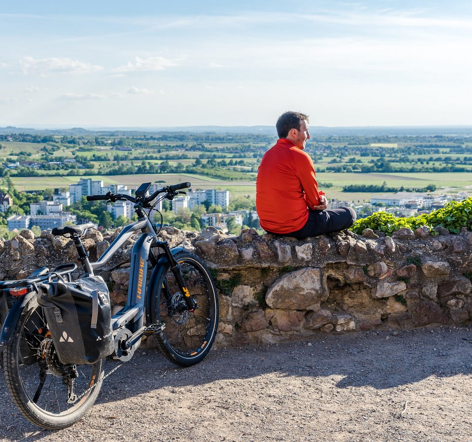 Mann in rotem Jacke sitzt auf Steinmauer mit E-Bike und Aussicht auf Stadt und Landschaft