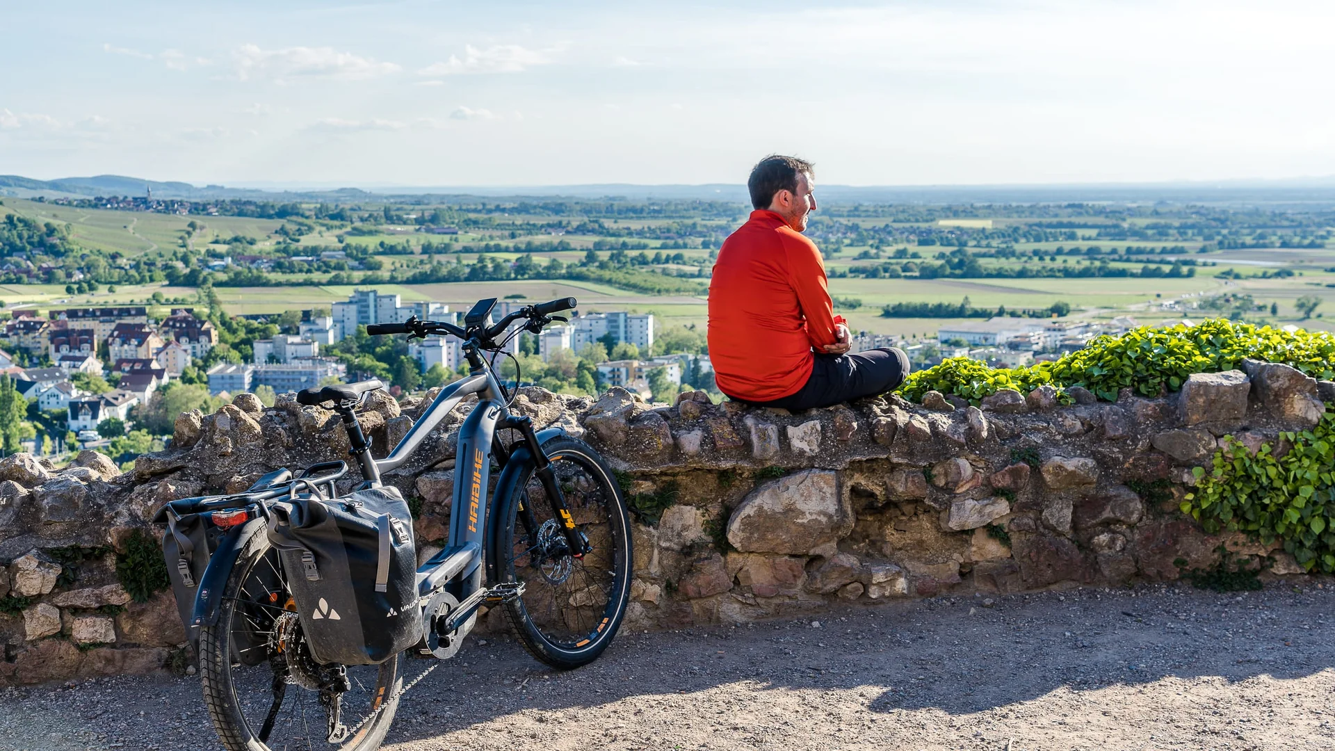 Man in rood jack zit op stenen muur met e-bike en uitzicht op stad en landschap