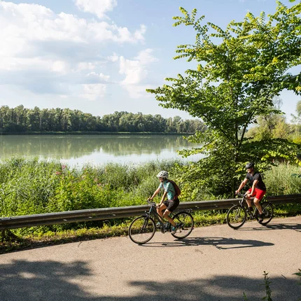 Zwei Radfahrer fahren auf einem Weg neben einem See im Grünen.