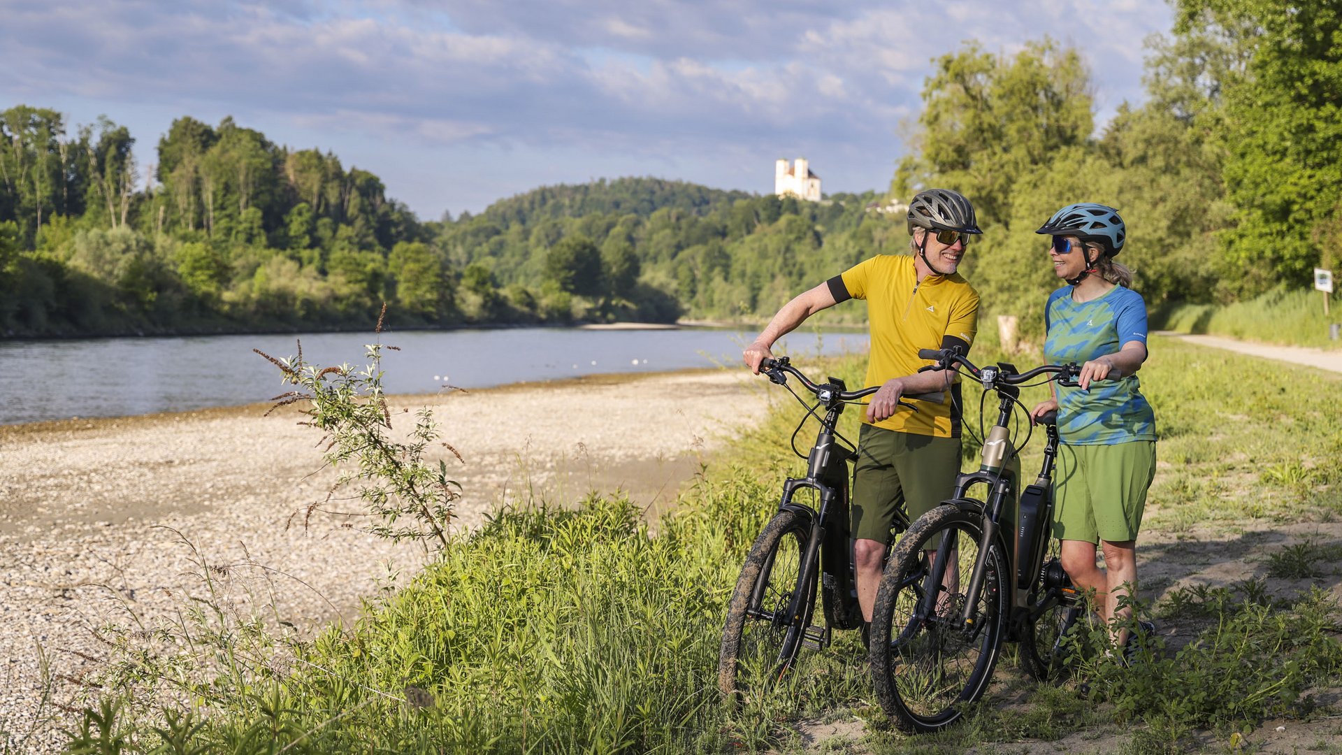 Two cyclists standing with bikes by riverbank with forested hills in background