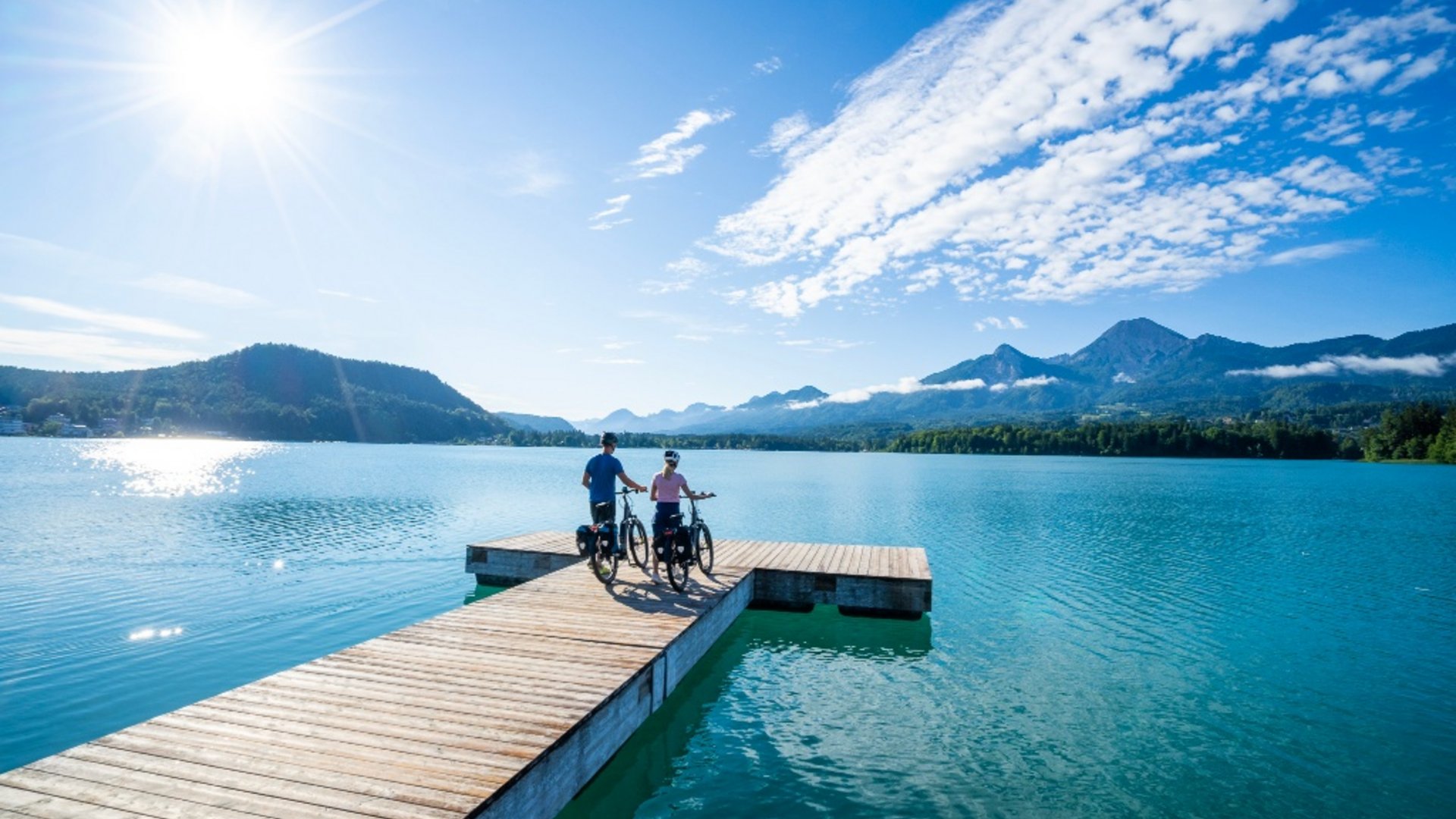 Two cyclists on a pier overlooking a lake and mountains on a sunny day
