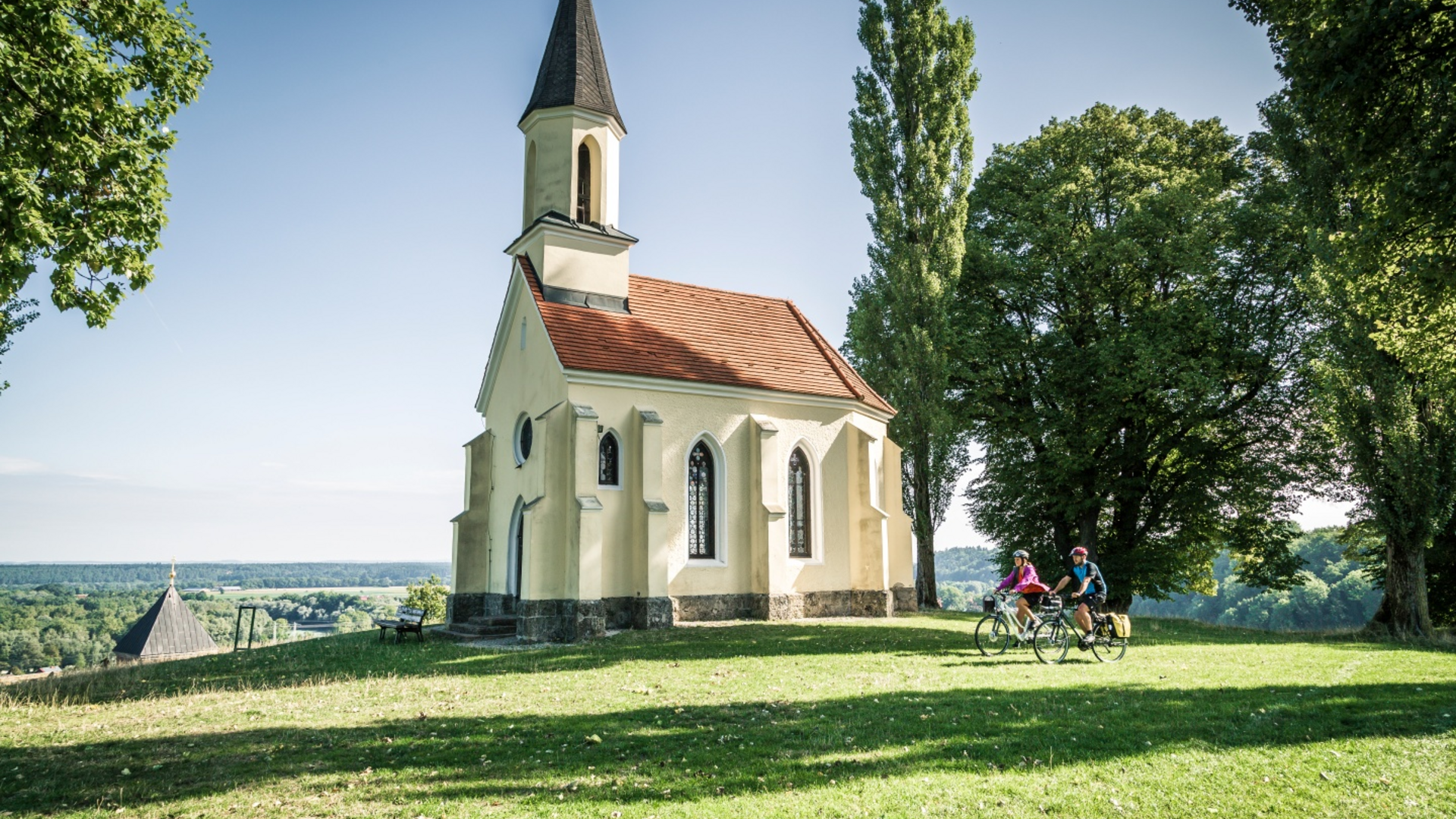 Small church on hill with two cyclists and trees on a sunny day