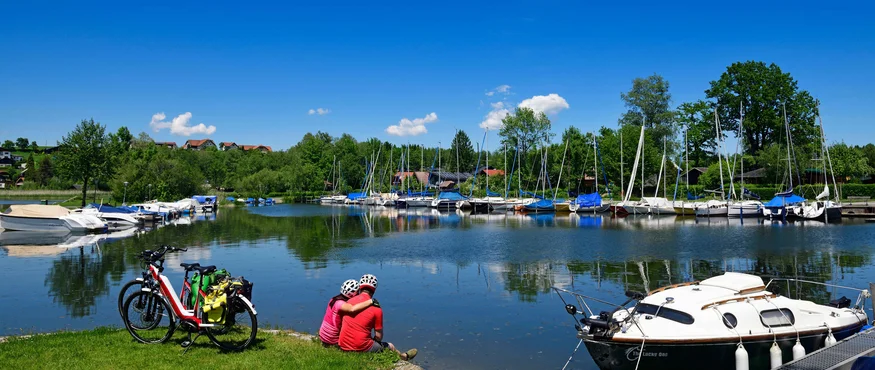 Couple with bikes sitting by lake shore near yachts on clear sunny day