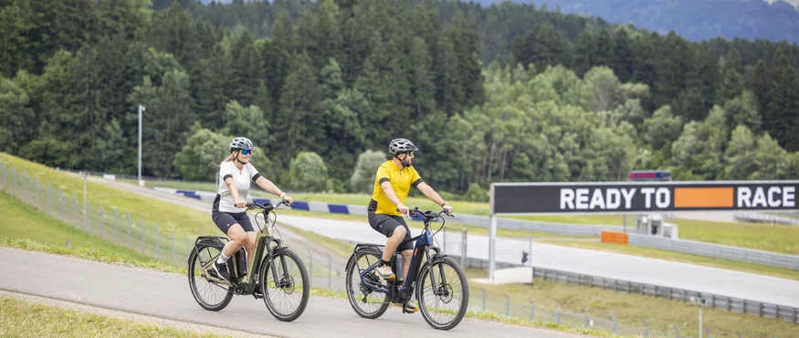 Zwei Radfahrer fahren auf einer Straße mit Wald und Bergen im Hintergrund