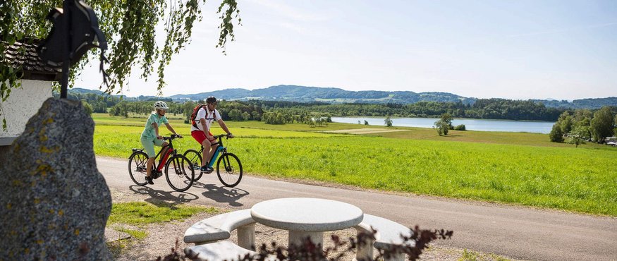 Two cyclists riding on a path near a lake and circular bench seating