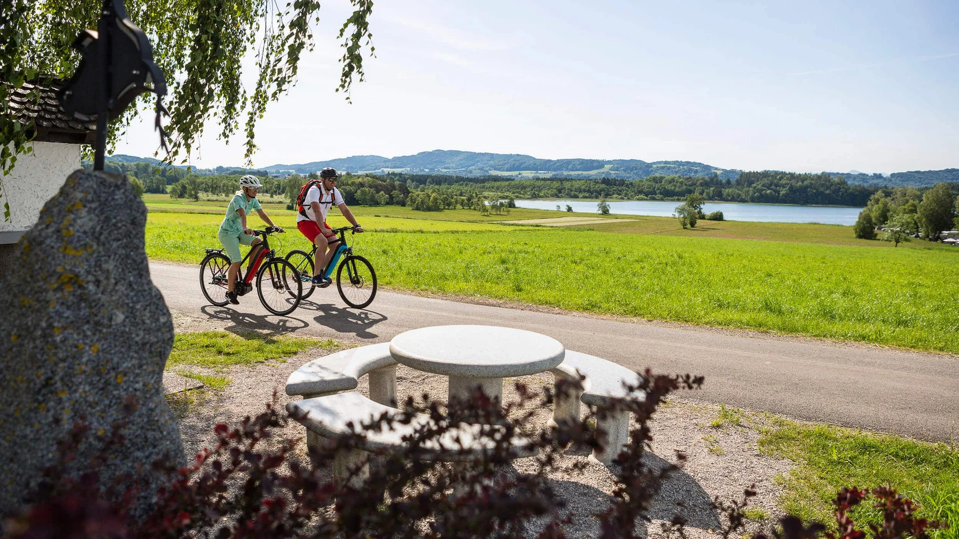 Zwei Fahrradfahrer fahren auf einem Weg neben einem See und einer Sitzgruppe