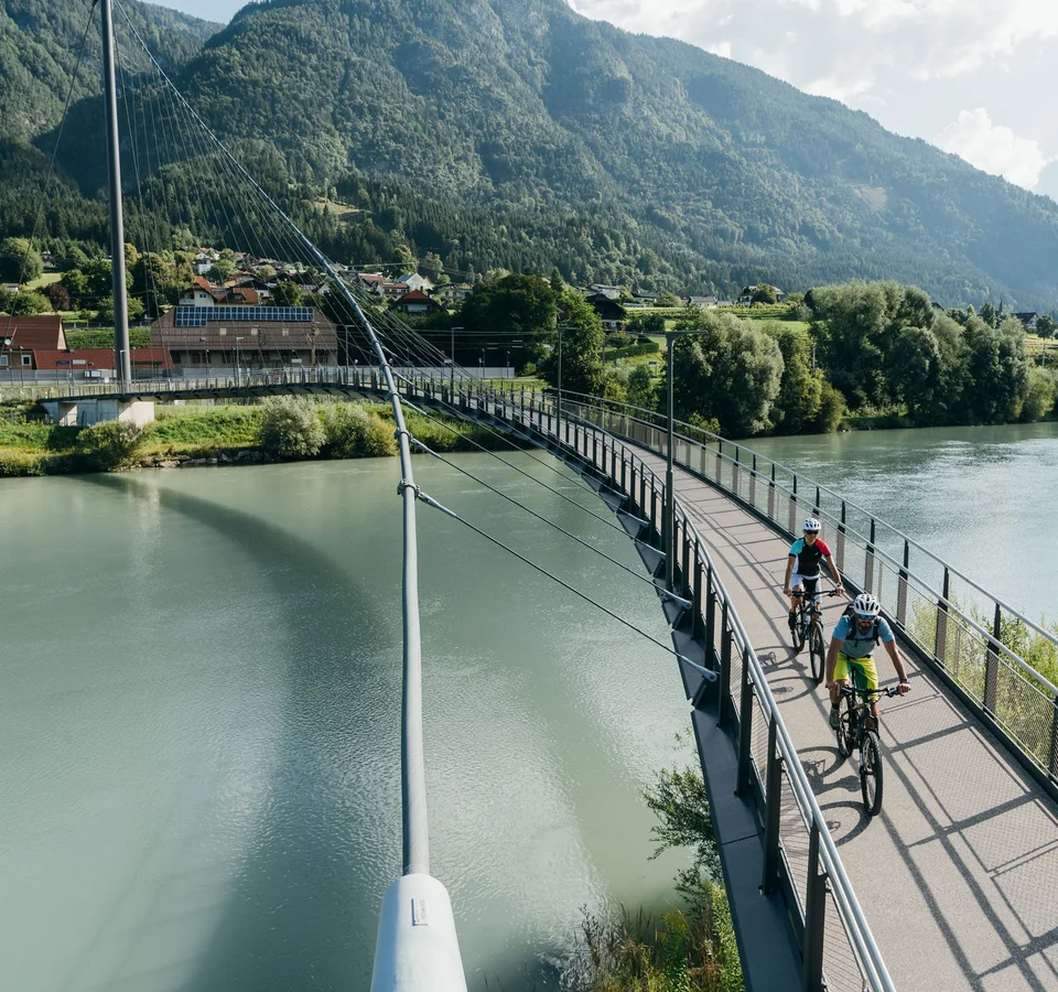 Zwei Radfahrer auf einer Fußgängerbrücke über einen Fluss in bergiger Landschaft