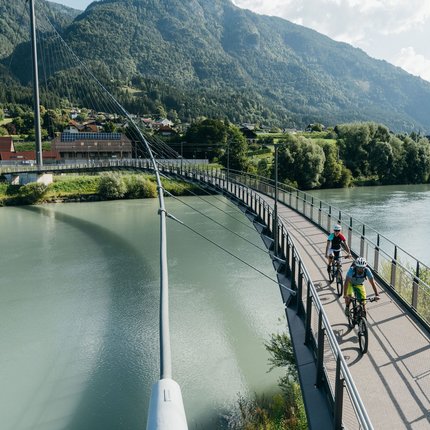 Two cyclists on a pedestrian bridge over a river in a mountainous area