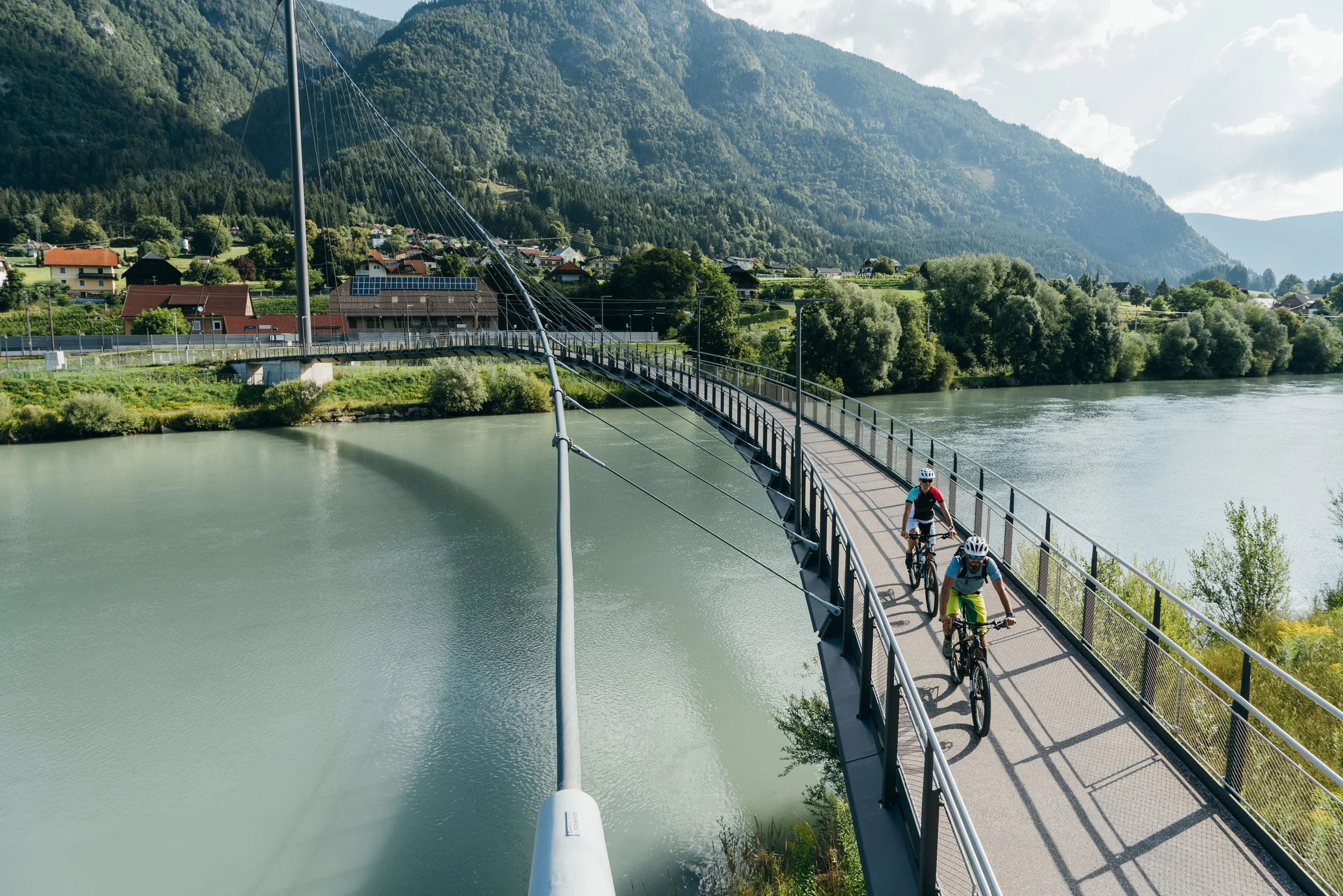 Zwei Radfahrer auf einer Fußgängerbrücke über einen Fluss in bergiger Landschaft