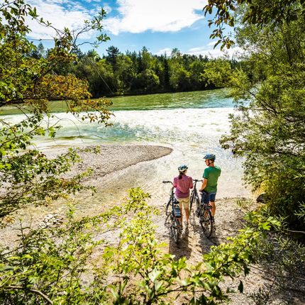 Two cyclists stand at riverbank surrounded by trees watching the water