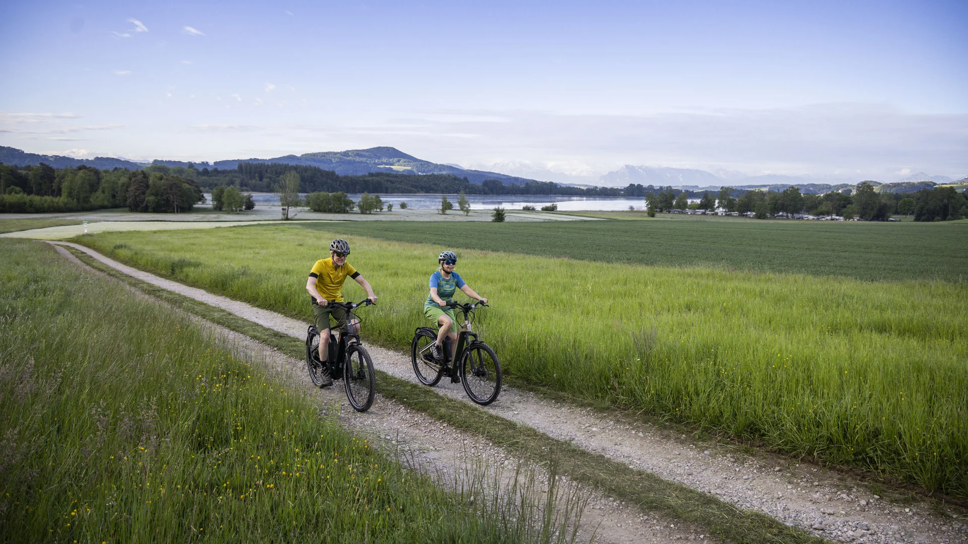 7 Quellen der Entschleunigung © Velontour - Heiko Mandl Zwei Radfahrer auf einem Feldweg neben Wiesen und See im Hintergrund