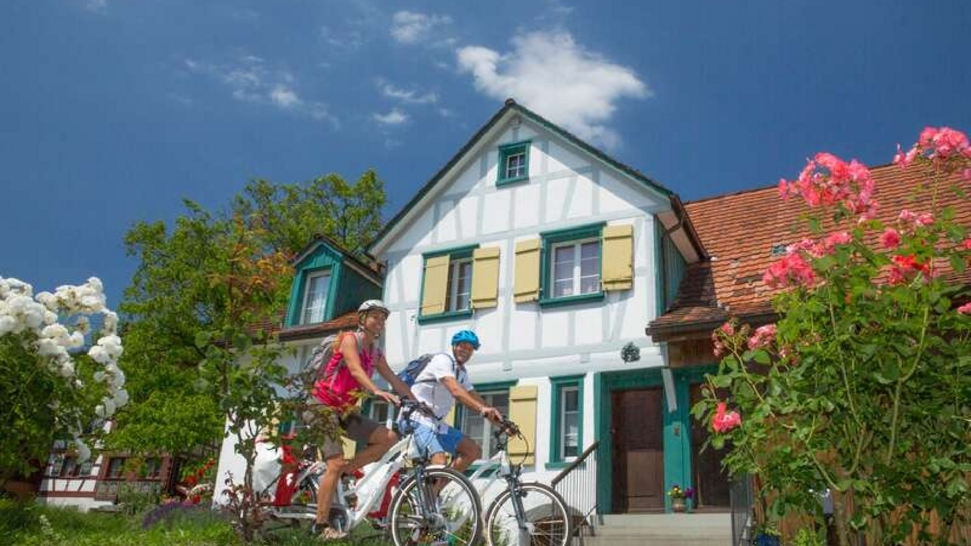Two cyclists in front of a traditional half-timbered house with garden and flowers