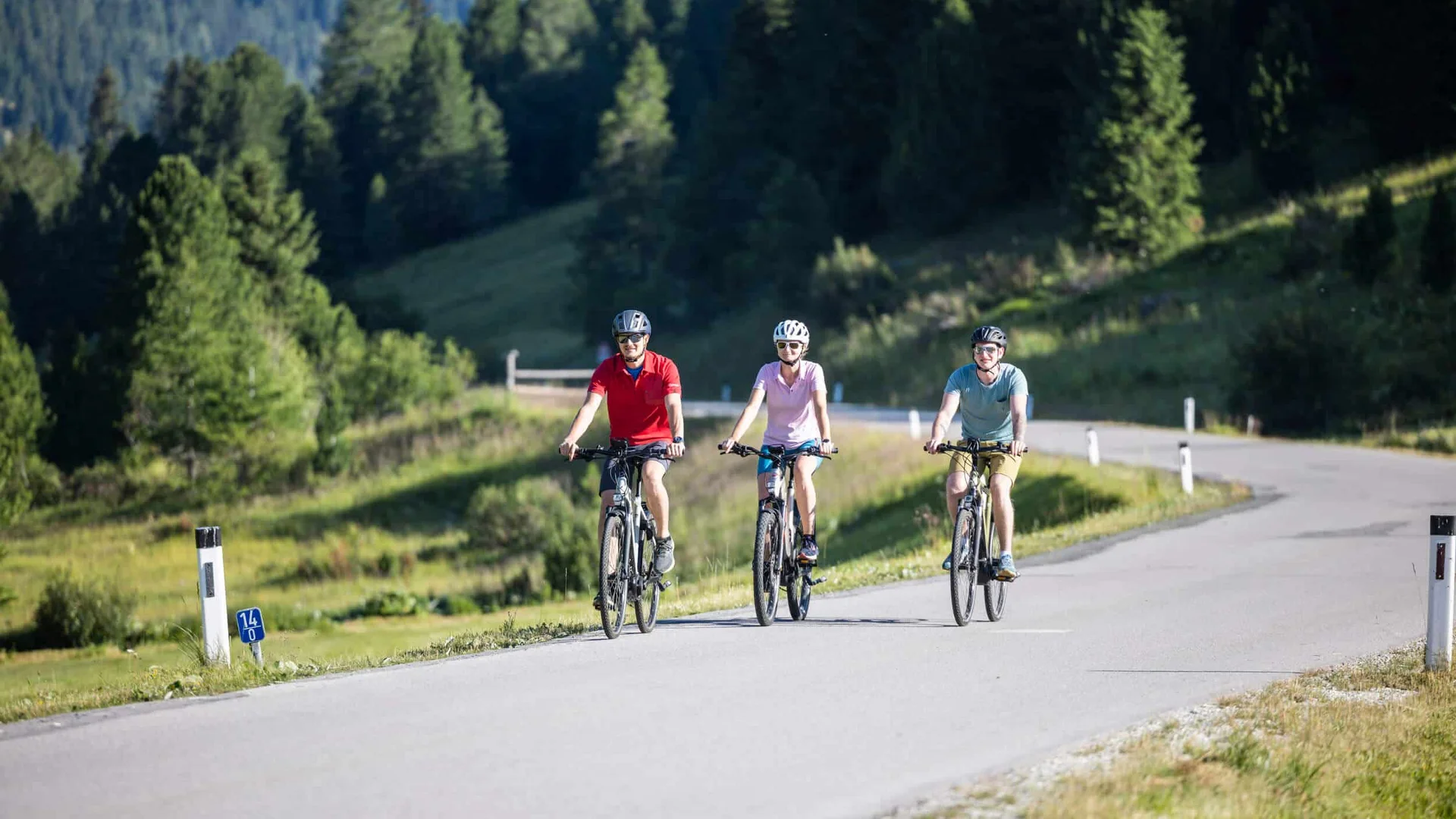 Drei Radfahrer fahren auf einer Landstraße durch grüne Berge