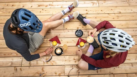 Zwei Radfahrer mit Helmen trinken Kaffee und essen Kuchen auf Holzterrasse
