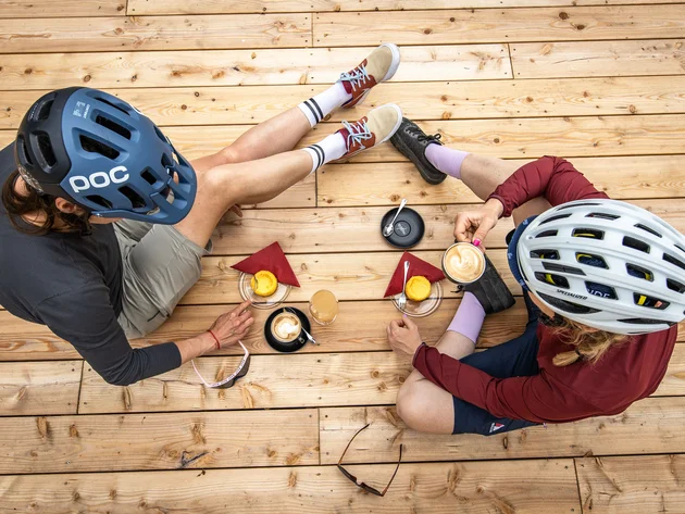 Zwei Radfahrer mit Helmen trinken Kaffee und essen Kuchen auf Holzterrasse