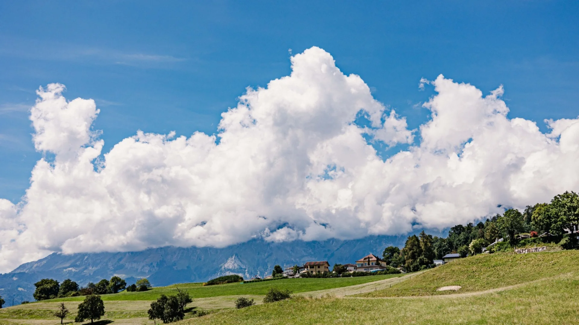 Grüne Wiese mit Häusern und Bergen unter einem blauen Himmel mit weißen Wolken