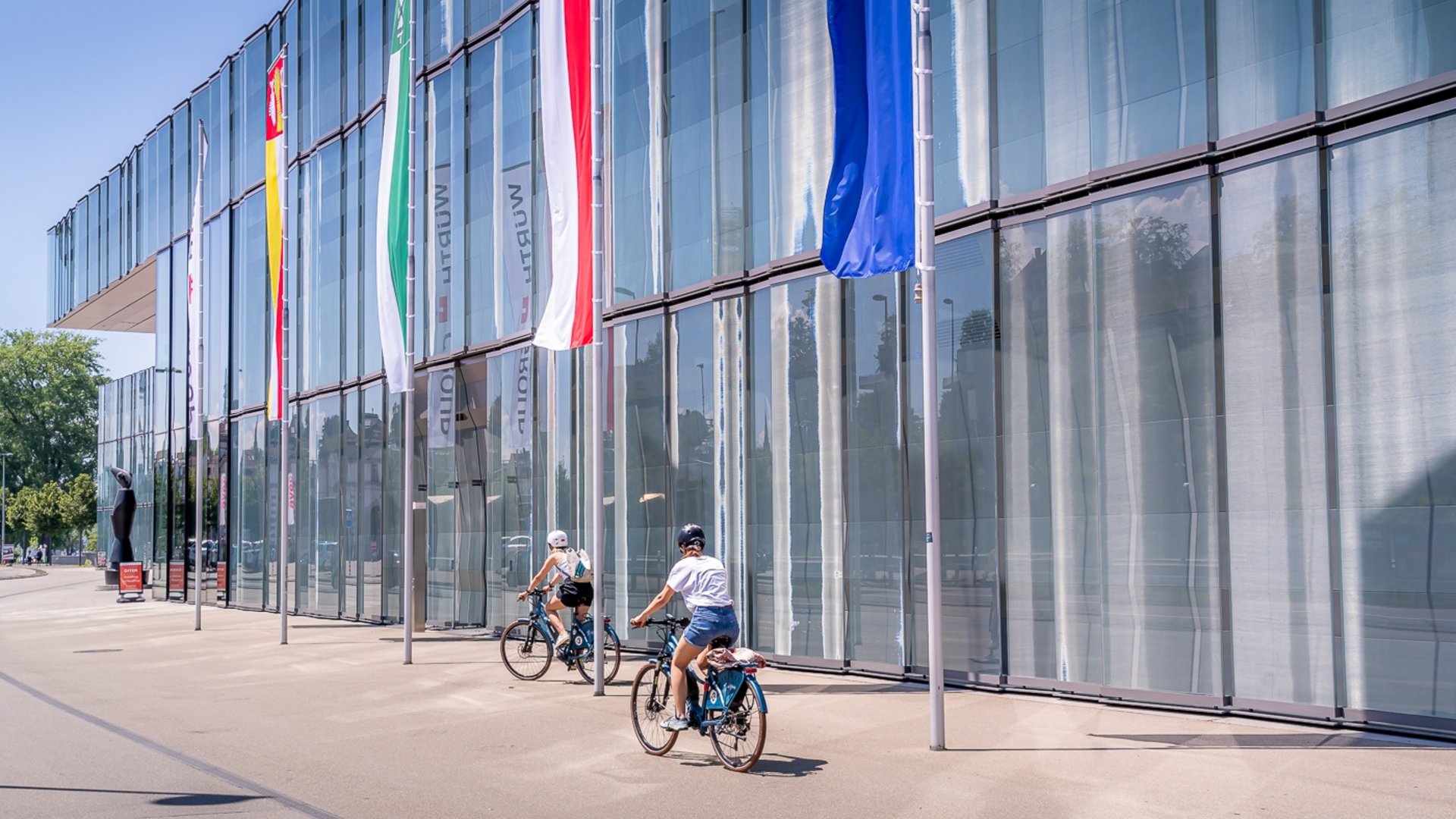 Two cyclists riding beside a glass building with flags