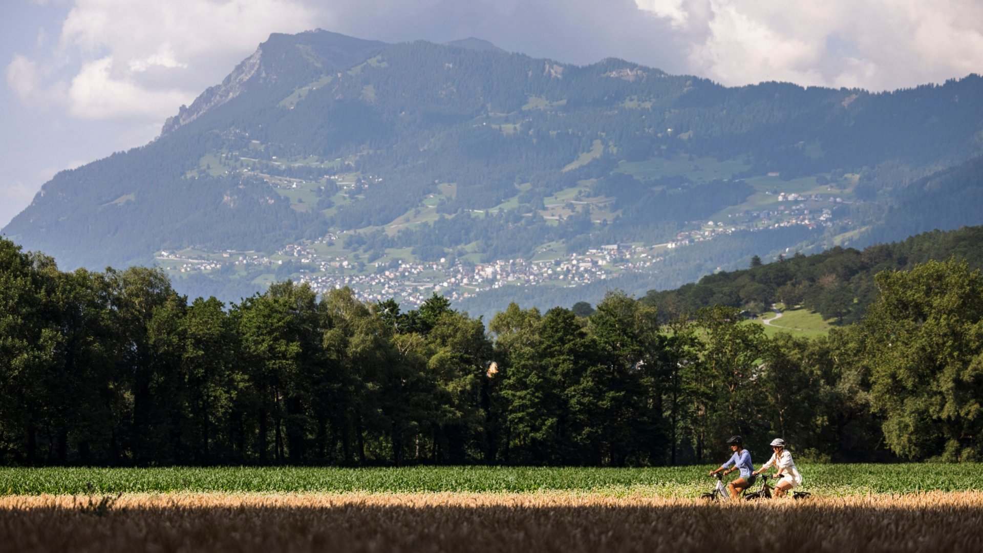 Two cyclists riding through green fields with a forested mountain in the background