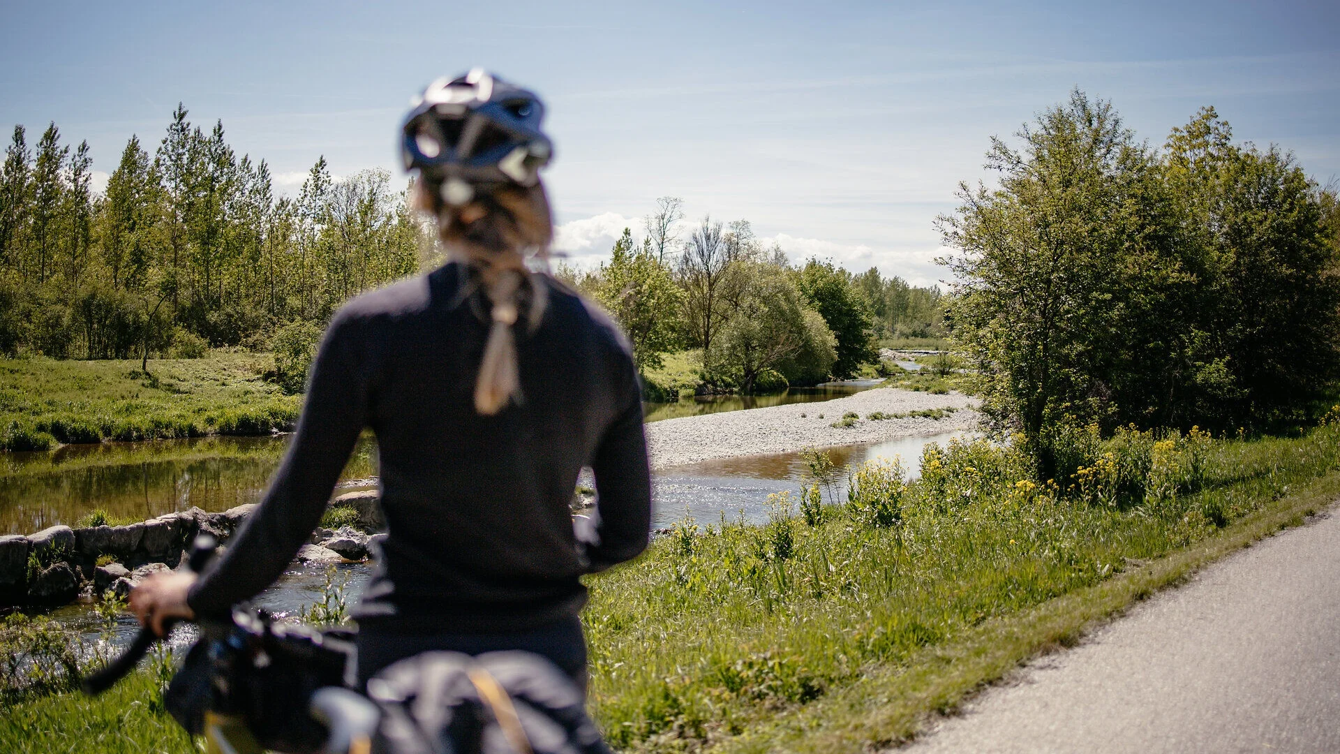 Tour-Detail Person mit Helm auf Fahrrad vor Fluss und grünen Bäumen bei klarem Himmel