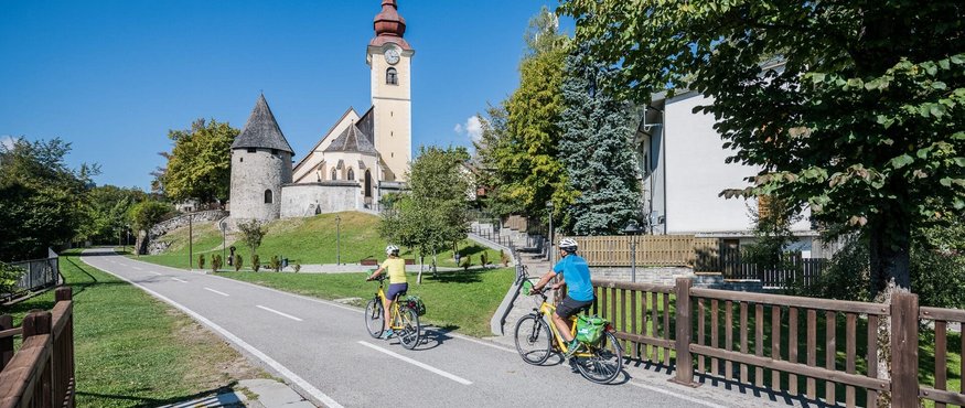 Two cyclists riding on a road near a church with tower on a sunny day