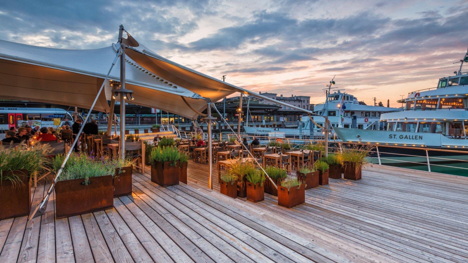 Evening terrace at harbor with tables, plants, and illuminated boats